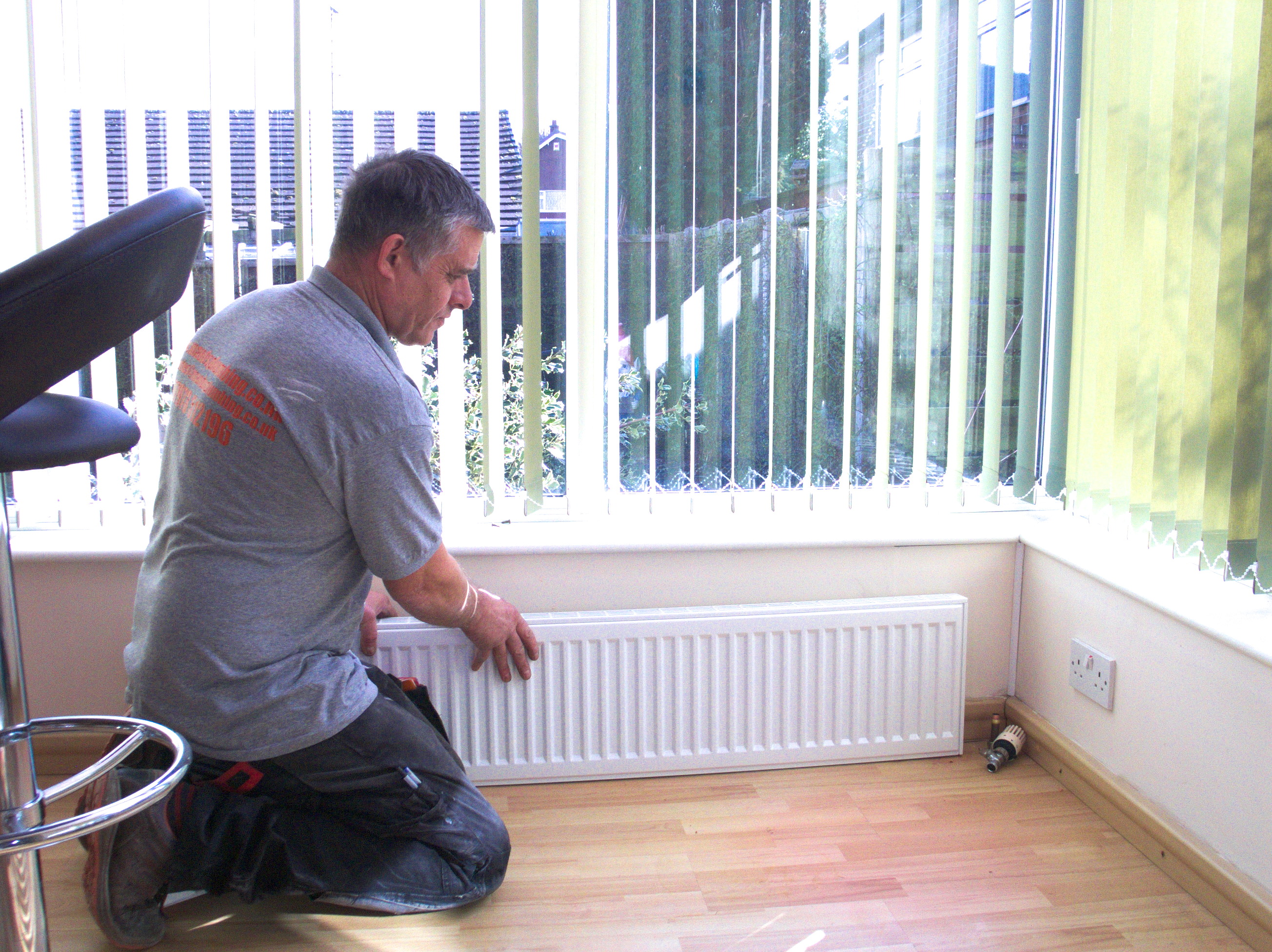 A man is kneeling on a wooden floor and installing a white radiator below a large window with vertical blinds.