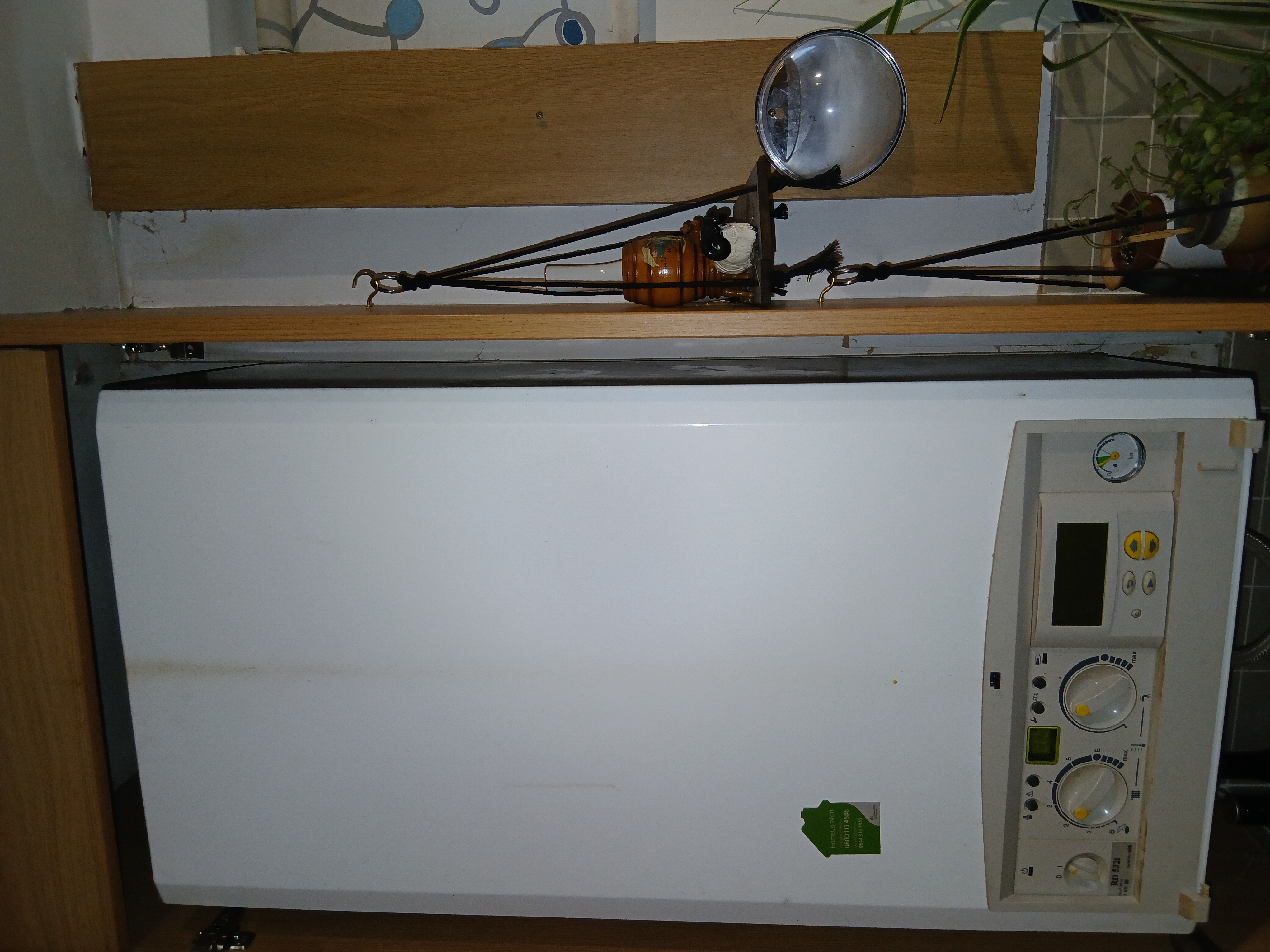 A white boiler with control dials and a digital display is installed in a wooden cabinet, with a small decorative shelf holding a plant and other items nearby.
